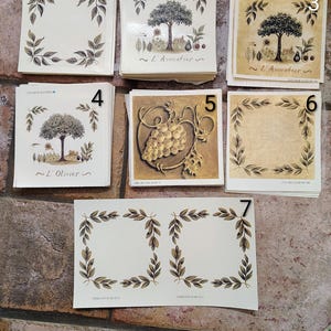 May include: A collection of seven square paper labels featuring botanical illustrations. Designs include olive branch wreaths, an avocado tree with the text L'Avocatier, an olive tree with the text L'Olivier, and a cluster of grapes in shades of green and gold.