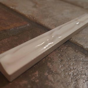 May include: A single, off-white ceramic tile trim piece with a glossy finish. The trim has a rectangular shape with a beveled edge and is set against a textured, multi-toned brown and gray surface. The tile is approximately 12 inches long.
