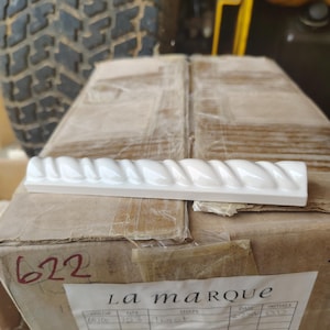 May include: A white ceramic tile with a rope-like design is displayed on a cardboard box. The box has the number "622" written on it in red marker. The label on the box reads "La Marque".
