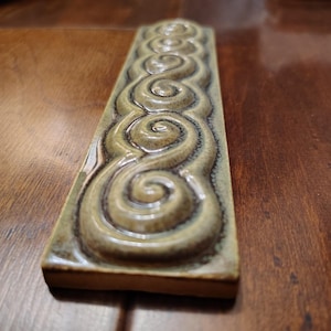 May include: A rectangular ceramic tile with a repeating spiral design. The tile has a glossy finish with shades of olive green and brown. The raised spiral pattern adds texture and visual interest to the tile.