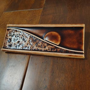 May include: A rectangular, brown ceramic dish with a textured, speckled design on one side and a smooth, circular shape on the other. The dish has a raised border and is set against a wooden surface.
