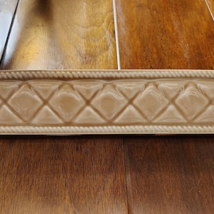 May include: A tan ceramic tile with a diamond pattern and rope-like trim. The tile is rectangular and appears to be a decorative element for a wall or other surface. The tile is approximately 12 inches long.