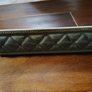 May include: A decorative trim piece with a quilted pattern in shades of green and gold. The trim has a rope-like border and is set against a wooden surface.