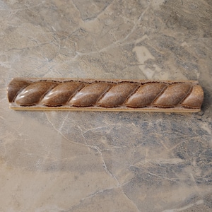 May include: A long, rectangular ceramic piece with a braided rope design. The ceramic is a light brown color with darker brown accents, and the surface has a glossy finish. The piece is likely a decorative element or trim.