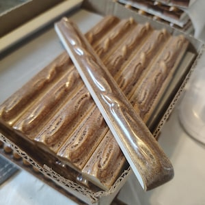 May include: A collection of brown ceramic tiles with a wood grain pattern, displayed in a cardboard box. The tiles have a glossy finish and a white edge. The tiles are approximately 12 inches long.