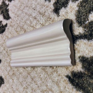 May include: A single piece of off-white ceramic molding with a glossy finish. The object features a curved, scalloped profile with multiple horizontal ridges running along its length, creating a decorative architectural trim piece.