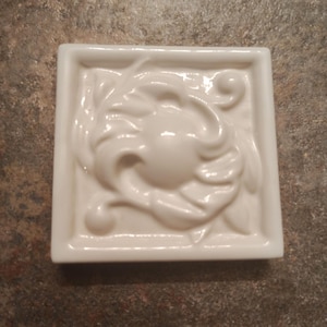 May include: A square, white ceramic tile with a raised floral design. The design features a central circular element surrounded by stylized leaves and curves. The tile has a glossy finish.