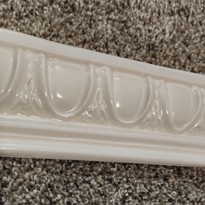 May include: Cream-colored decorative molding with a repeating, ornate design. The molding has a glossy finish and is rectangular in shape. The design features a series of curved, symmetrical elements. This architectural detail is likely used for interior design.
