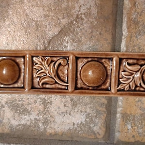 May include: A rectangular, brown ceramic tile with four square sections. Two sections feature a raised, circular design, while the other two showcase a detailed leaf and vine pattern. The tile has a glossy finish and is likely for home decor.