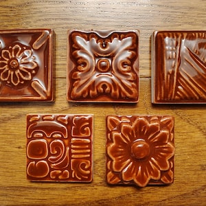 May include: Five square, brown ceramic tiles with various embossed designs. The tiles feature floral, geometric, and abstract patterns. The tiles are arranged on a wooden surface, showcasing their glossy finish and intricate details.