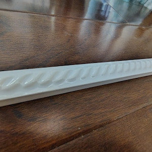 May include: A white, rectangular decorative molding with a repeating wave pattern along one edge. The molding is made of a solid material and is displayed on a wooden surface.