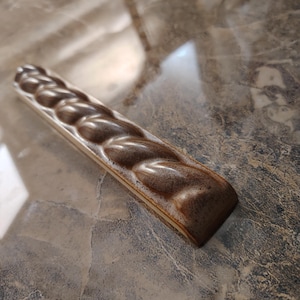 May include: A brown ceramic soap dish with a braided design. The rectangular dish has a raised, textured surface with a series of rounded, interconnected shapes. The color is a warm, earthy brown with lighter highlights.