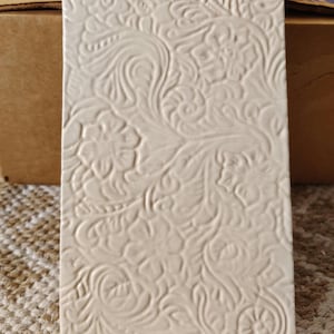 May include: A rectangular, off-white ceramic tile with an embossed floral pattern. The design features swirling vines and stylized flowers, creating a textured surface. The tile is likely for home decor or a craft project.
