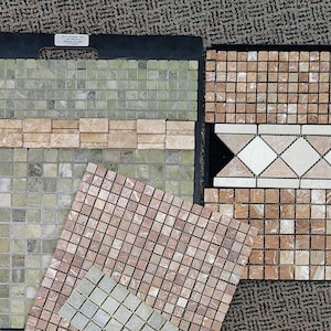 May include: A collection of stone mosaic tile samples arranged on display boards. The set includes square tiles in shades of sage green, tan, and pink, alongside a decorative border piece featuring a repeating diamond pattern in cream and beige.