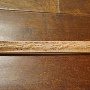 May include: A rectangular, light brown wooden decorative trim piece with a carved, wavy design running along its length. The trim has a glossy finish and is likely for home decor or craft projects.