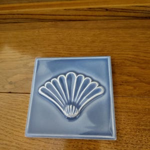 May include: A square, light blue ceramic tile with a raised, white seashell design in the center. The tile is set on a wooden surface. The seashell design has a fan-like shape.