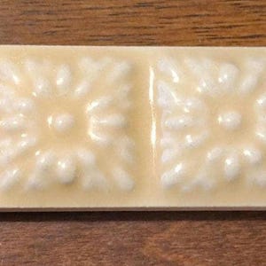 May include: A rectangular, off-white ceramic tile strip featuring six square tiles. Each tile has a raised, floral design with a central dot and surrounding petals. The tiles are arranged in a row, creating a decorative element.