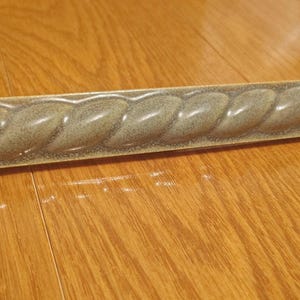 May include: A decorative trim piece with a rope-like design. The trim is a muted green color with a speckled texture and a slightly curved shape. It is lying on a wooden surface.