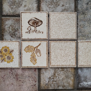 May include: A set of five square ceramic tiles with a speckled cream finish. Three tiles feature brown line art illustrations: one with a mushroom, one with yellow flowers, and one with an acorn and oak leaves. Two tiles are plain and unadorned.