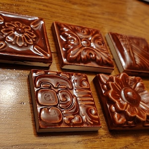 May include: Five square, brown ceramic tiles with raised floral and geometric designs. The tiles are arranged on a wooden surface, showcasing their glossy finish and intricate patterns. Each tile has a white border.