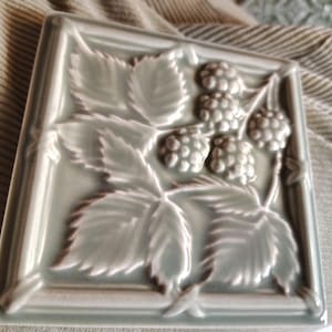 May include: Square, light green ceramic tile with a raised relief design of raspberries and leaves. The tile is framed with a simple border. The design is detailed and the tile has a matte finish.