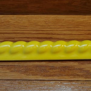 May include: A bright yellow ceramic object with a horizontal, elongated shape featuring a repeating twisted rope pattern along the top edge and a smooth, flat base. The surface has a glossy, reflective finish that catches the light.
