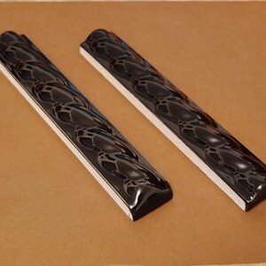 May include: Two long, rectangular ceramic tiles featuring a glossy, deep black finish with a raised, repeating wave-like pattern along the center. Each tile has a smooth, rounded top surface and a flat, white base, showcasing a reflective texture.