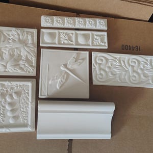 Peut inclure: Un ensemble de six carreaux en plâtre blanc, de formes carrées et rectangulaires, présentant divers motifs en relief tels qu'une libellule, des fleurs, des branches fruitières et des bordures décoratives avec une finition mate.