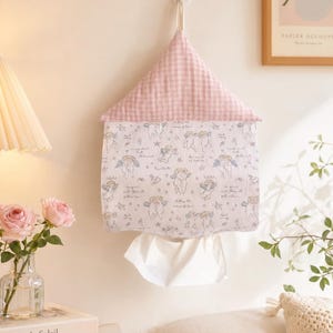 May include: A pink and white gingham tissue box cover shaped like a house, hanging on a wall. The lower portion features a white fabric with a pattern of cherubs and stars. White tissues are visible at the bottom.