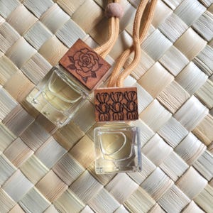 Pode incluir: Duas pequenas garrafas de vidro transparente cheias de um líquido amarelo claro, cada uma encimada por um pequeno quadrado de madeira gravada. Um quadrado apresenta um desenho de rosa, o outro um padrão repetitivo. As garrafas penduram de um cordão castanho sobre um fundo tecido.