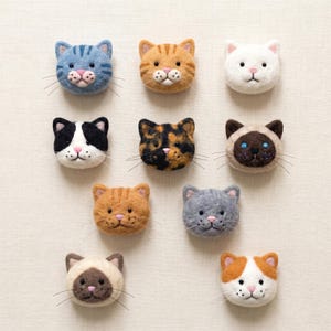 May include: A collection of eleven felted cat head ornaments in various colors and patterns. The cats include tabby, calico, Siamese, and solid colors like white and gray. Each cat has a pink nose and whiskers.