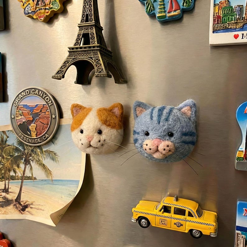 May include: Two felt cat head magnets, one orange and white, the other blue with stripes, are displayed on a refrigerator. A yellow taxi cab magnet and a miniature Eiffel Tower are also visible, along with other travel-themed magnets.
