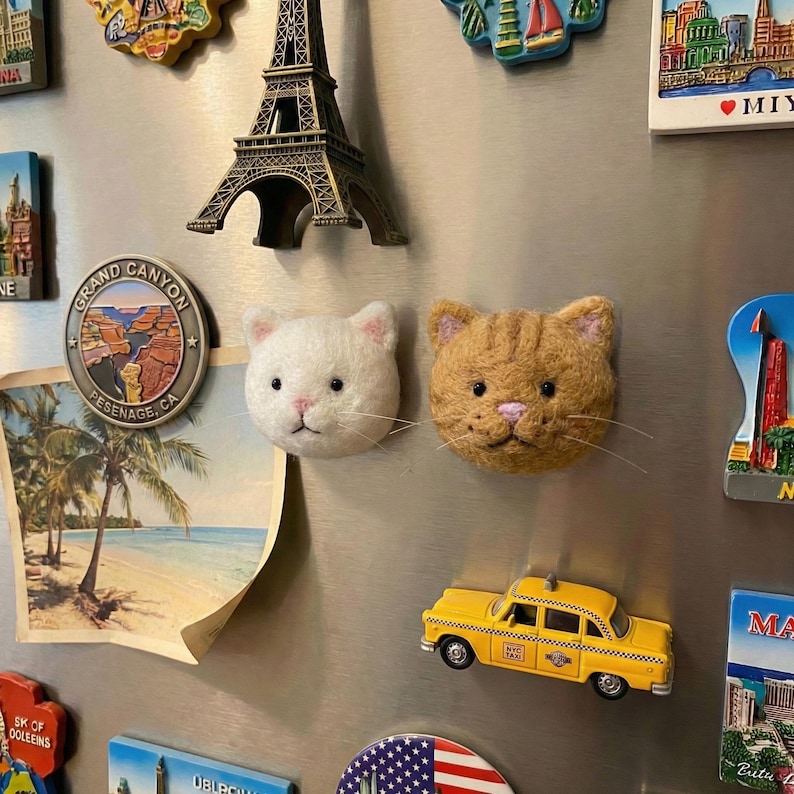 May include: A collection of refrigerator magnets, including a miniature Eiffel Tower, a Grand Canyon souvenir, and a yellow taxi cab. Two felt cat head magnets are also present, one white and one orange.