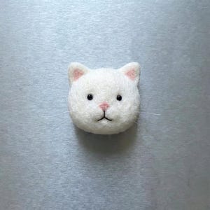 May include: A white felt cat head with pink inner ears, a pink nose, and black eyes. The cat has thin, white whiskers. The cat is centered on a light gray background.