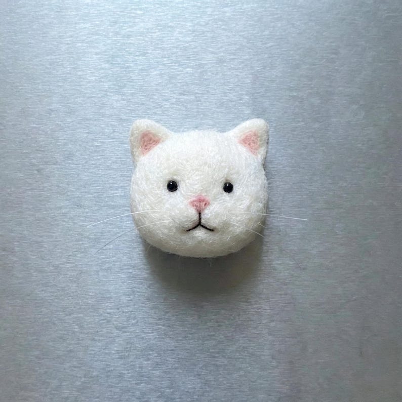 May include: A white felt cat head with pink inner ears, a pink nose, and black eyes. The cat has thin, white whiskers. The cat is centered on a light gray background.