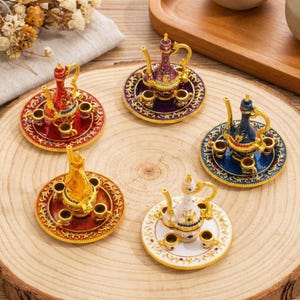 May include: Miniature decorative tea sets in red, purple, blue, gold, and white. Each set includes a small teapot and several tiny teacups on a matching saucer. The sets are intricately detailed with gold accents and colorful enamel.