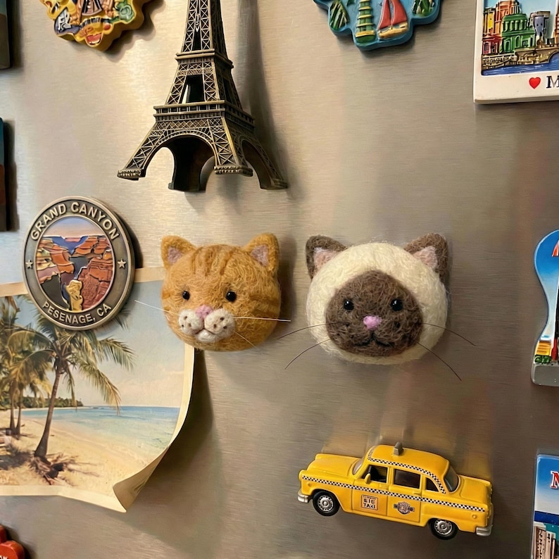 May include: A collection of refrigerator magnets, including a miniature Eiffel Tower, a Grand Canyon souvenir, and a yellow taxi cab. Two cat-shaped magnets are also present, one orange tabby and one Siamese cat.