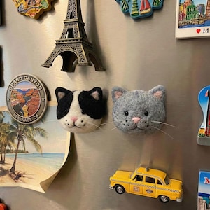 May include: A collection of decorative magnets on a silver refrigerator. The magnets include a black and white cat face, a gray cat face, a yellow taxi cab, a miniature Eiffel Tower, and a Grand Canyon souvenir.