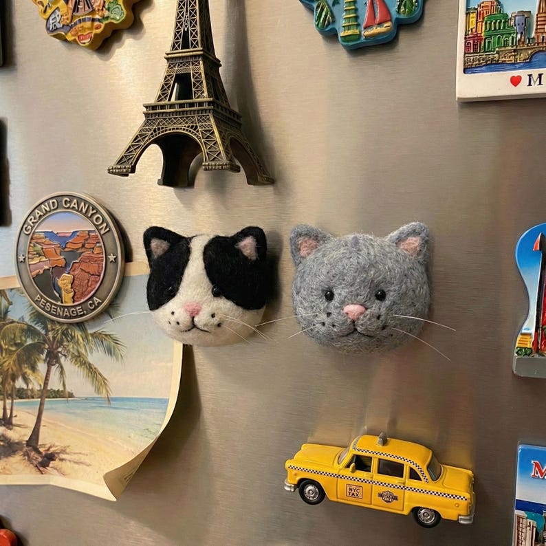 May include: A collection of decorative magnets on a silver refrigerator. The magnets include a black and white cat face, a gray cat face, a yellow taxi cab, a miniature Eiffel Tower, and a Grand Canyon souvenir.