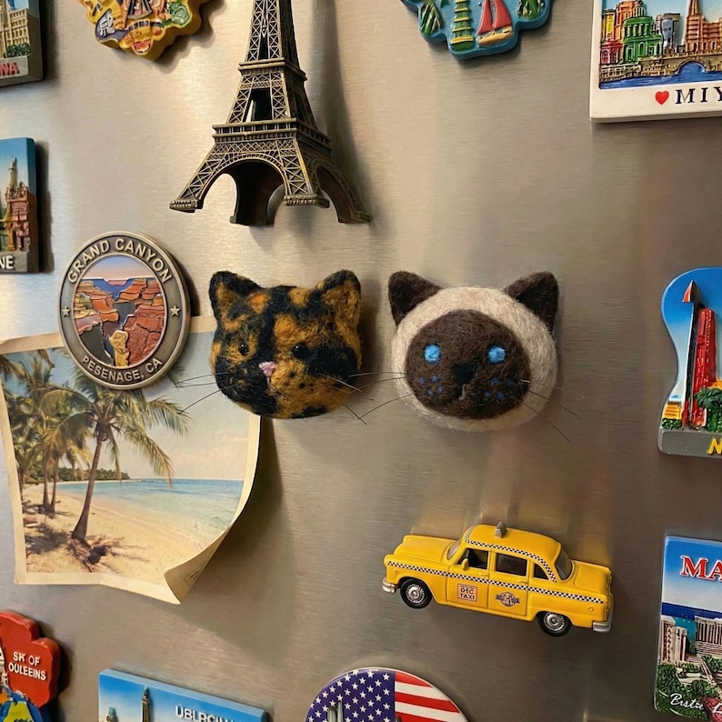 May include: A collection of refrigerator magnets, including two cat-shaped magnets, a yellow taxi cab, a miniature Eiffel Tower, and a Grand Canyon souvenir. The cat magnets are brown and tan, and the taxi cab has the word "TAXI" on the roof.