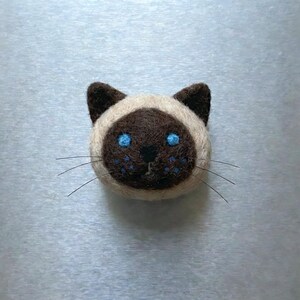 May include: A handcrafted Siamese cat face made of felt. The cat has a brown face and ears, with blue eyes and whiskers. The cat's face is surrounded by a cream-colored felt.