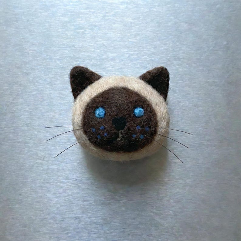 May include: A handcrafted Siamese cat face made of felt. The cat has a brown face and ears, with blue eyes and whiskers. The cat's face is surrounded by a cream-colored felt.