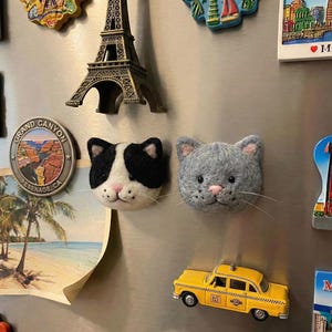 Puede incluir: Una colección de imanes para nevera, que incluye dos imanes de fieltro con forma de gato en blanco y negro y gris. Otros imanes incluyen un taxi amarillo, una Torre Eiffel en miniatura y un recuerdo del Gran Cañón.