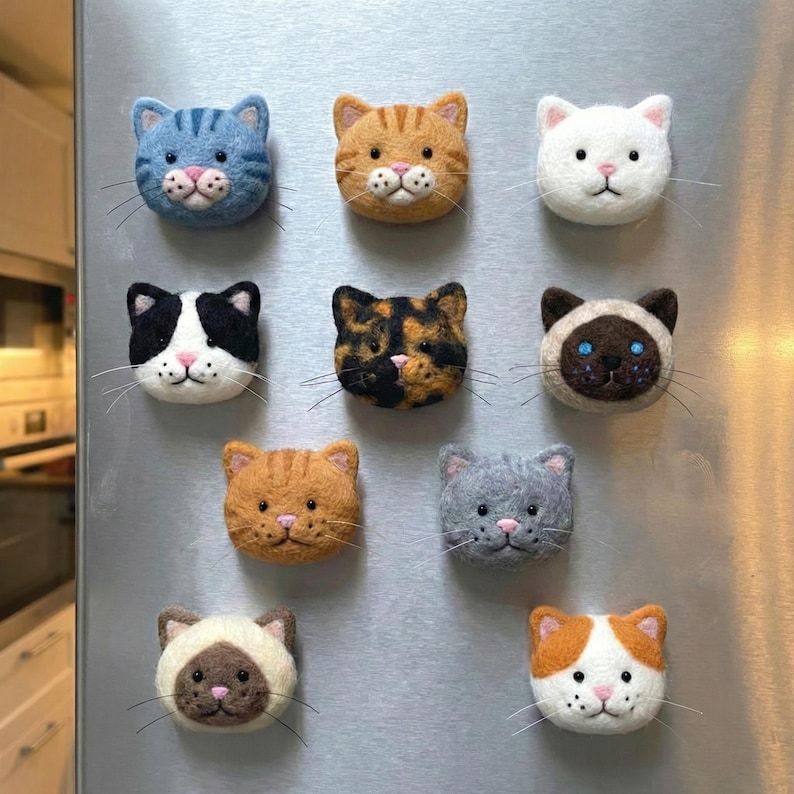 May include: A collection of handmade cat-shaped magnets in various colors and patterns. The magnets feature detailed facial features, including whiskers and pink noses. The cats are made of felt and are arranged on a metallic surface.