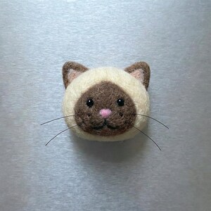 May include: A handcrafted Siamese cat face made of felt. The cat has a cream-colored face with brown markings, a pink nose, and black whiskers. The cat's eyes are black and it has a sweet expression. This felt cat face is a charming decorative item.