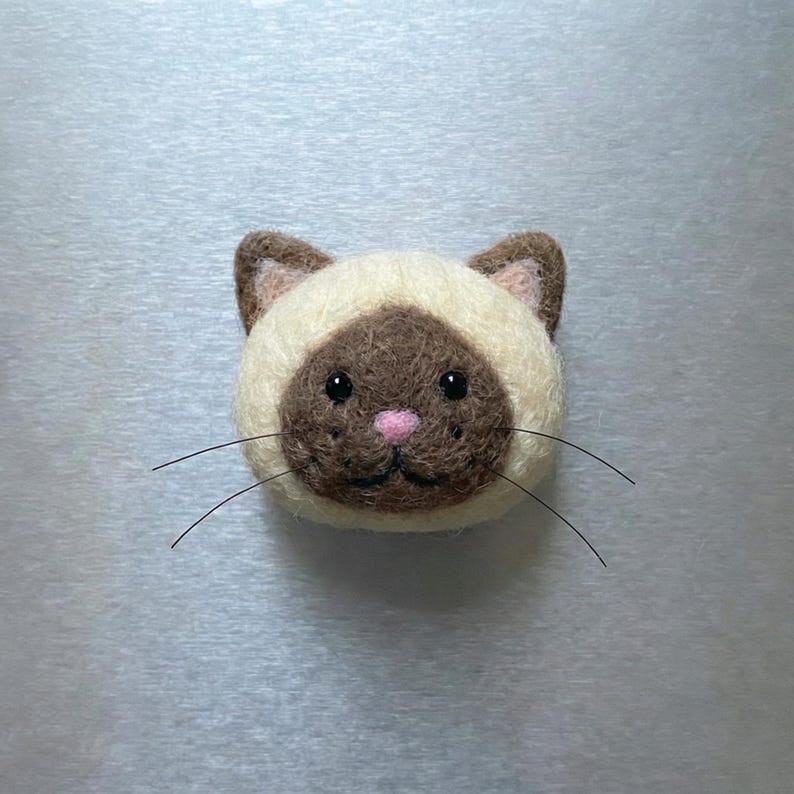 May include: A handcrafted Siamese cat face made of felt. The cat has a cream-colored face with brown markings, a pink nose, and black whiskers. The cat's eyes are black and it has a sweet expression. This felt cat face is a charming decorative item.