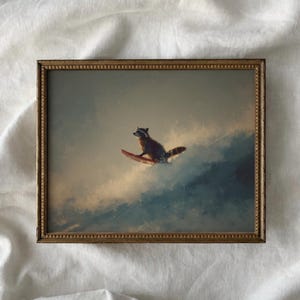 May include: A framed print depicting a raccoon surfing on a wave. The raccoon is brown and grey, riding a red surfboard. The background shows a cloudy sky in shades of blue and grey. The frame is gold.