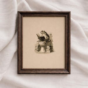 May include: A framed wall art print featuring a black and white ink illustration of a frog sitting on a pedestal while reading an open book, surrounded by stacks of books, enclosed in a dark brown wooden frame with a textured finish.