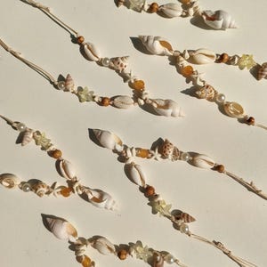 Puede incluir: Varios collares de conchas marinas con diversos tipos de conchas, perlas y cuentas de madera. Los collares están ensartados en cordones beige y dispuestos diagonalmente sobre un fondo blanco. Algunos collares incluyen pequeñas conchas en forma de estrella y cuentas de color ámbar.