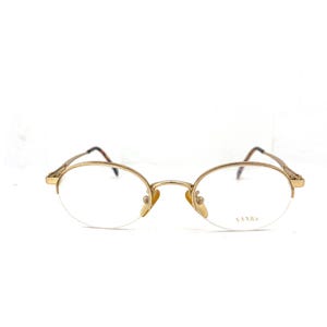 以下が含まれることがあります： ゴールドトーンの楕円形メガネ、ハーフリムデザイン。クリアレンズと繊細なフレームが特徴です。右レンズにはブランド名「VANNA」が見えます。テンプルは濃い茶色です。